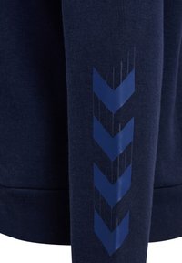 Mörkblå sweatshirt med mjukt tyg, ribbade muddar och ett glänsande blått chevronmönster på ärmen. Minimalistisk design och texturaccenter.