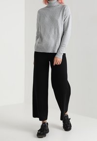 Grijze coltrui van zacht materiaal, gecombineerd met zwarte wijde broek en zwarte sneakers, staand tegen een neutrale achtergrond.