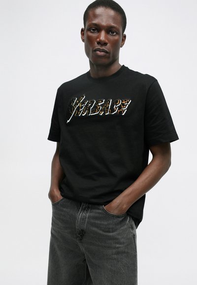 Camiseta preta de algodão com uma gráfica texturizada e multicolorida "Versare" ao centro. Combinada com jeans cinzentos.