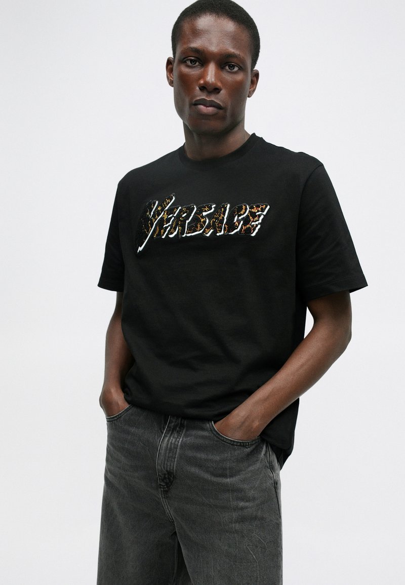 T-shirt en coton noir avec un graphique texturé et multicolore "Versare" au centre. Associé à un jean en denim gris.