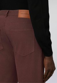 Burgunderfarbene Denim-Jeans mit glatter Textur, ausgestattet mit zwei Gesäßtaschen und einem braunen Lederlabel am Bund. Schwarzes Oberteil teilweise sichtbar.