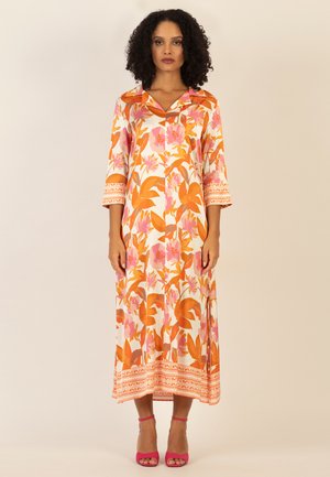 Frau mit lockigem Haar, die ein langes, florales Kleid in Orange und Rosa trägt, mit entspannt hängenden Armen, kombiniert mit pinken offenen Schuhen mit Absatz.