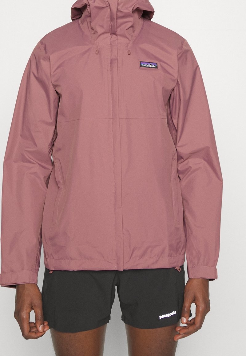 Homme portant une veste à capuche rose poudré avec fermeture éclair à l'avant et logo de la marque sur la poitrine gauche, associée à un short noir avec logo de la marque sur la cuisse gauche.
