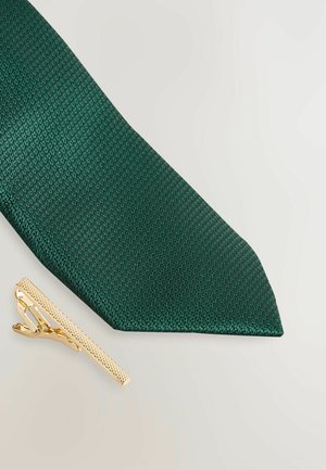 Cravatta verde testurizzata con punta, abbinata a una clip per cravatta dorata con superficie rigata e meccanismo di chiusura.