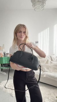 Frau, die eine schwarze Lederhandtasche mit doppelten Griffen und einem Schulterriemen in einem hellen, modernen Wohnzimmer hält.