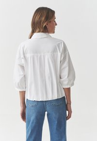 Camicia bianca oversize con colletto, maniche a palloncino corte e dettaglio di piega nella parte posteriore. Abbinata a jeans di denim chiaro.
