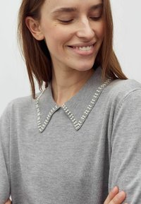 Fiatal nő mosolyogva lefelé néz, szürke pulóvert visel, melynek nyakrésze gyöngyökkel díszített, valamint apró arany fülbevalót.