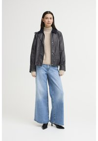 Svart croppad jacka med krage, lagd över en beige stickad tröja. Ljusblå vida jeans och svarta spetsiga klackar bärs.