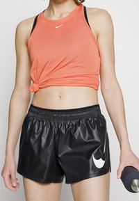 Top corto senza maniche di color corallo con dettaglio a nodo, abbinato a pantaloni corti neri lucidi con il logo bianco della Nike sul lato.
