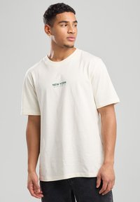 T-shirt en coton couleur crème avec des manches courtes et un col rond, portant l'inscription "NEW YORK" en vert sur la poitrine.