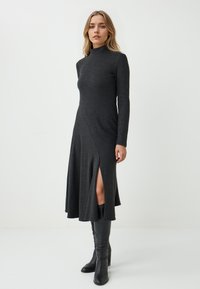 Abito grigio a maniche lunghe con colletto alto, spacco laterale e orlo svasato. Realizzato in tessuto morbido; abbinato a stivali neri al ginocchio.