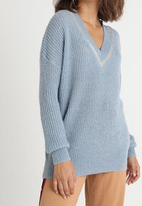 Ljusblå stickad tröja med V-hals och silverdetaljer, med en avslappnad passform samt ribbade manschetter och nederkant. Strukturerad tyg synlig.