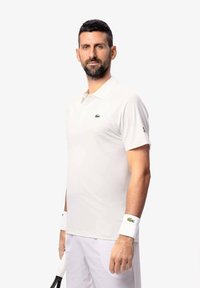 Hvid polo t-shirt med korte ærmer, prydet med et grønt Lacoste-logo. Båret med hvide håndledsband og holder en tennisketsjer.