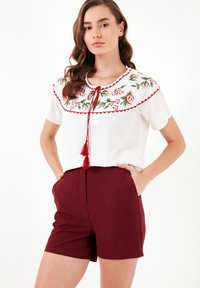 Witte top met bloemenborduursel in roze en groen, rode afwerking en een striksluiting. Gecombineerd met kastanjebruine shorts met zijzakken.