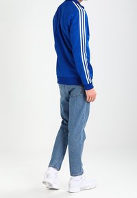 Blauwe sportieve jas met witte strepen langs de mouwen, gecombineerd met lichtblauwe jeans en witte sneakers. Glad materiaal, comfortabele pasvorm.