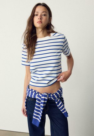 Giovane donna con lunghi capelli castani che indossa una maglietta a righe bianche e blu e jeans scuri con un maglione a righe abbinato legato intorno alla vita.