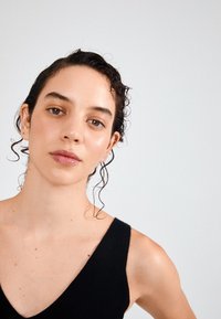 Juodas išilginis tankiniukas su V formos iškirpte, turintis plonas petnešėles. Modelio plaukai šlapi, garbanojati, o oda glotni, su minimaliais aksesuarais.