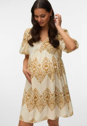 Tehotná žena s dlhými tmavými vlasmi, oblečená v krémových šatách s horčicovými ozdobnými vzormi, usmievajúca sa a dotýkajúca sa vlasov a brucha.