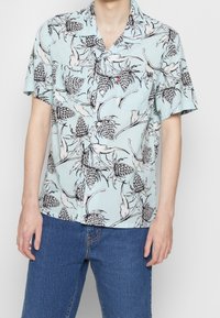 Camicia azzurro chiaro a maniche corte con motivo floreale nero e forme simili a pigne. Il tessuto sembra leggero e liscio.