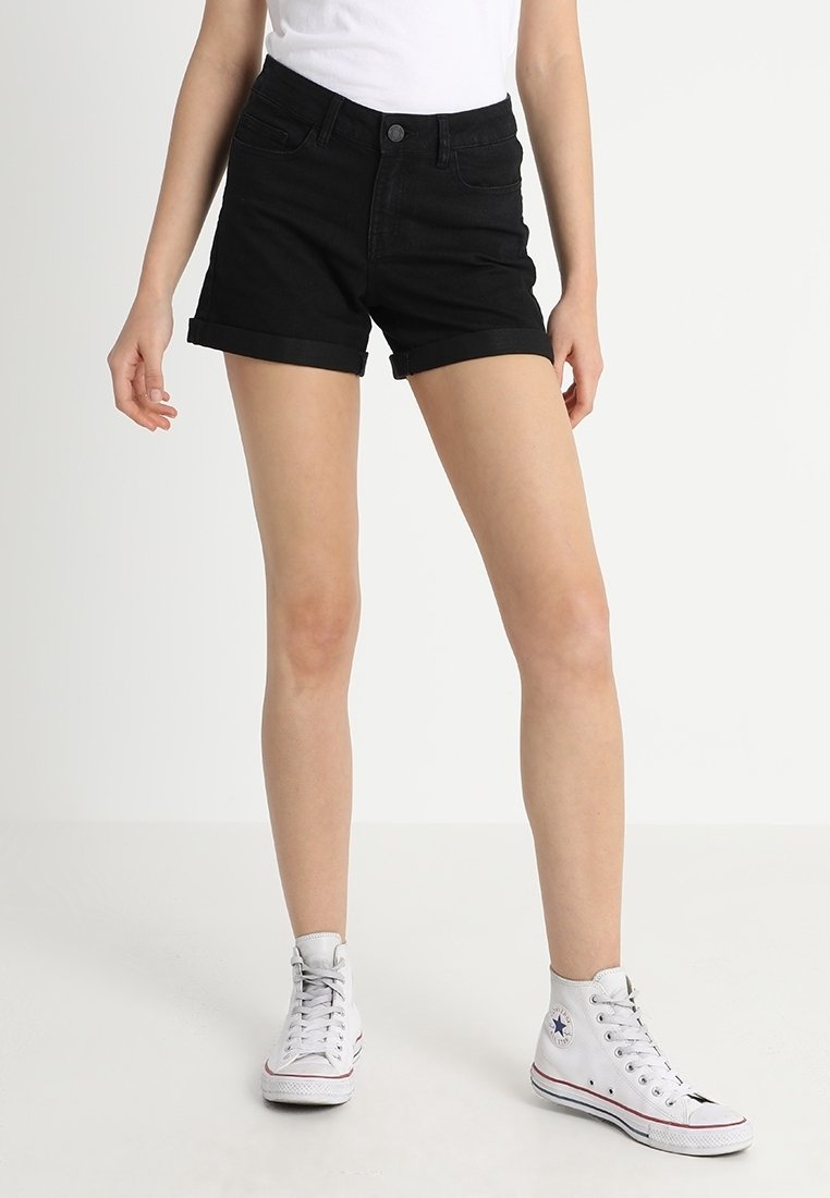 Femme portant un short noir à ourlet roulé, un t-shirt blanc et des baskets montantes blanches, debout devant un fond uni.