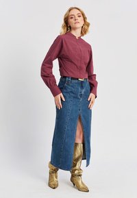 Donna che indossa un top lungo maniche color marrone, una gonna in denim blu a vita alta con uno spacco frontale e stivali metallici dorati al ginocchio, in posa con sicurezza.