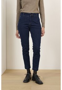 Jean en denim bleu foncé à coupe ajustée, taille haute et ourlets retroussés, associé à des chaussures habillées noires et des chaussettes noires texturées.