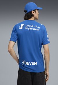 Blå atletisk skjorte med korte ærmer, med hvid tekst for "Riyad Bank" og "SEVEN" på ryggen, parret med en matchende blå kasket.