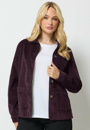Femme blonde portant une chemise blanche et une veste en velours côtelé violet foncé avec des boutons et des poches avant, debout devant un fond uni.