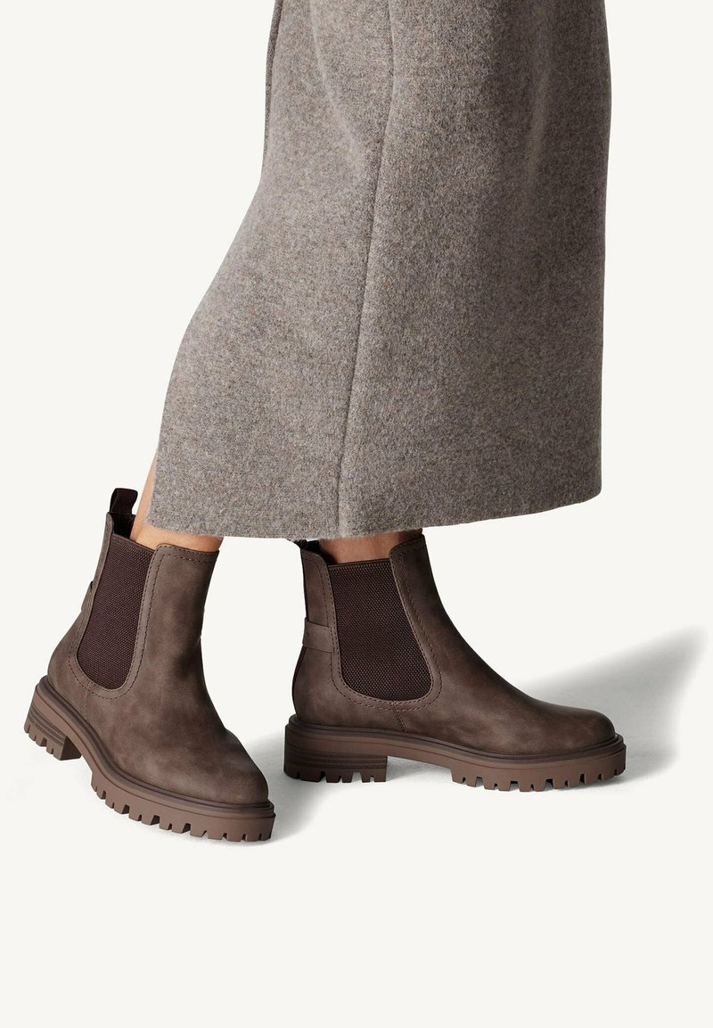 Bottes Chelsea en cuir marron avec semelles épaisses en caoutchouc, portées avec une longue jupe en laine grise sur un fond uni.