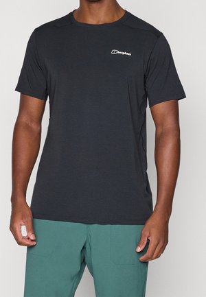 Mann trägt schwarzes Berghaus T-Shirt mit kurzen Ärmeln und grüne Hose, steht vor einfachem hellem Hintergrund.