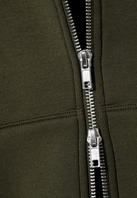 Κοντινή λήψη ασημένιων διπλών φερμουάρ σε ενδυμασία από σκούρο πράσινο ύφασμα με μερικώς ανοικ τό φερμουάρ.