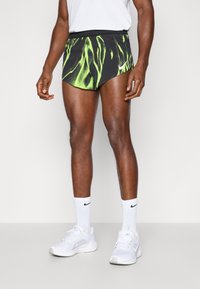 Nike melni šorti ar dzīvīgu zaļu abstraktu rakstu, elastīga jostasvieta un kontrastējoša šuvuma, savienoti ar baltām kedām un īsām zeķēm.