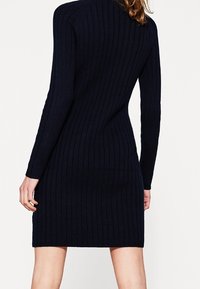 Robe en maille côtelée bleu marine avec des manches longues, silhouette ajustée, décolleté rond et texture subtile, atteignant la longueur mi-cuisse à l'arrière.