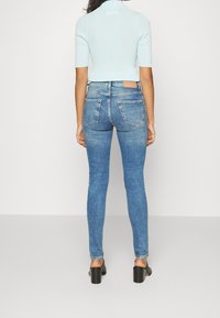 Úzké džíny ve středně modré denim s vybledlým povrchem, s zadními kapsami a hnědou koženou nášivkou na pase. Nosené s světle modrým topem.
