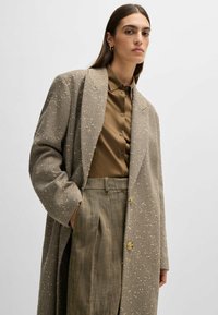 Oversized beige herringbone coat with textured accents, featuring large buttons, paired with a brown button-up shirt and patterned trousers.