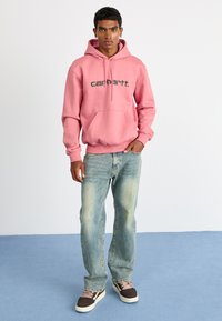 Pink hoodie with a front pocket and black logo, paired with faded blue jeans and brown sneakers with light laces on a blue floor.