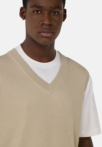Gilet en tricot côtelé beige avec un décolleté en V profond, porté par-dessus un T-shirt blanc. La texture du tissu est fine et lisse, mettant en valeur son design superposé.