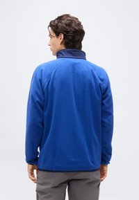 Νέος άνδρας με την πλάτη γυρισμένη φοράει ένα βασιλικό μπλε φλις μπουφάν με ψηλό γιακά και γκρι παντελόνι, σε ένα απλό λευκό φόντο.