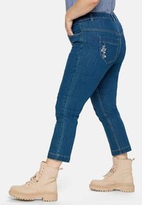 Mörkblå denimjeans med medelhög midja, avsmalnande ben och blommigt broderi på bakfickan. Kombinerat med beige snörstövlar.