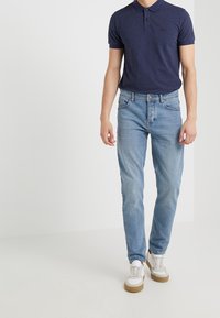 Muži v námořnickém modrém polo tričku, světle modrých džínech a bílých teniskách s gumovými podrážkami stojí před bílým pozadím.