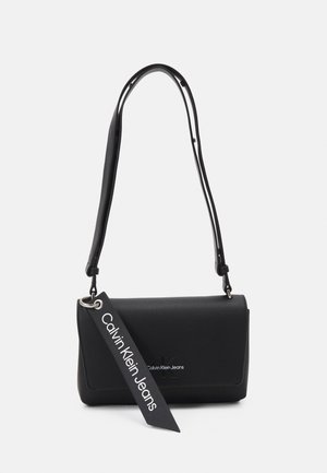 Bolso de hombro negro de Calvin Klein Jeans con correa ajustable y etiqueta de la marca colgando de la solapa frontal sobre un fondo blanco.