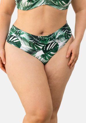 Frau trägt grün-weiß bedruckte Bikinihose mit tropischem Blättermuster und passendem Oberteil, steht mit entspannt an den Seiten liegenden Händen.