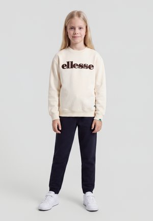 Jeune fille aux longs cheveux blonds portant un sweat ellesse crème, un pantalon noir et des baskets blanches, debout devant un fond gris uni.
