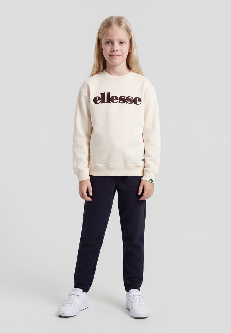 Jeune fille aux longs cheveux blonds portant un sweat ellesse crème, un pantalon noir et des baskets blanches, debout devant un fond gris uni.