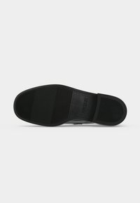 Semelle de chaussure noire avec des motifs texturés d’adhérence et la taille 37 marquée près de la zone du talon sur un fond gris clair uni.