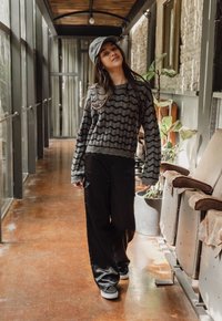 Grijze gebreide trui met golfpatroon, ruimzittende zwarte pantalons en donkere sneakers. De setting heeft een houten vloer en groenvoorzieningen op de achtergrond.