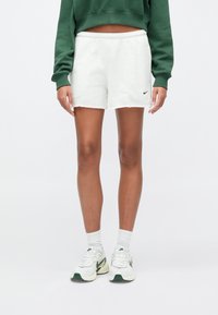 Άτομο που φοράει λευκό σορτς Nike, λευκές κάλτσες μέχρι τον αστράγαλο, λευκά και πράσινα αθλητικά παπούτσια Nike και ένα κοντό πράσινο φούτερ, στέκεται μπροστά σε λευκό φόντο.