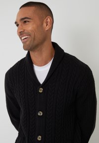 Cárdigan negro de punto trenzado con cuello de chal, que cuenta con cuatro botones redondos y un patrón texturizado. Llevado sobre una camiseta blanca.