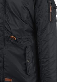 Veste noire rembourrée avec un motif matelassé, une poche zippée avec des accents marron et un ajustement à la taille par cordon de serrage.