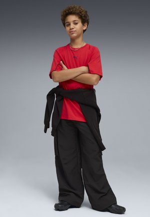 Chłopiec o kręconych włosach stoi pewnie, z założonymi na piersi rękami, ubrany w czerwoną koszulę, czarne spodnie, czarne buty oraz czarną kurtkę zawiązaną wokół talii.